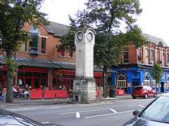 A stone tower on a street of brick buildings which include a public house and a café with tables and chairs in front of it.