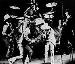 A group of men performing on a stage.  One is playing a violin, three are playing guitars, and one is playing drums.  The drummer and two of the guitarists are wearing cowboy hats.
