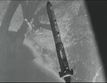 View from a high-altitude aircraft, looking straight down and showing a bomb that has just being released. On the ground can be seen a box-like building next to water