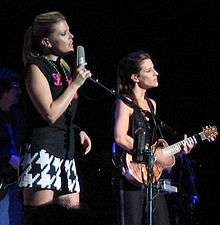 Two young women performing on a stage, one singing into a microphone and the other playing a guitar