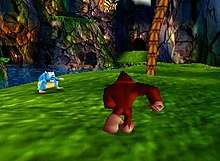 A brown gorilla runs across a green expanse of blurry green. A palm tree grows in the back right corner and a dark jungle background shows in the distance.