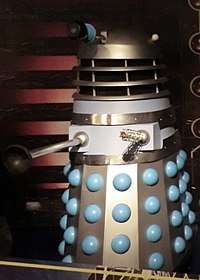 An original Dalek, coloured mostly silver and grey, with blue balls protruding from the skirt.