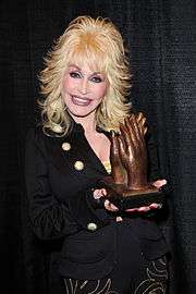 A blonde woman wearing dark clothing, holding a trophy in the shape of hands applauding