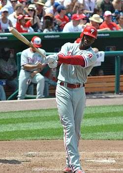 A dark-skinned young man wearing a red baseball helmet and a gray baseball jersey and pants with red trim swinging a baseball bat