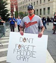 A mustached Caucasian man wearing a black baseball hat and a gray baseball jersey with "Baltimore" in orange script trimmed with black across the chest holds from the top, at waist level, a handmade sign with "Don't Forget Freddie Gray" written on it in black. Behind him are city streets with tall buildings and other people walking around