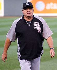 A man with a black cap and black and gray jersey with "Sox" written on each in white walking on a baseball field