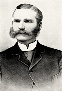 Formal half-length portrait of a middle-aged man with long sideburns, a full mustache, and neatly combed hair parted in the middle. He is wearing a dark suit and a white collar.