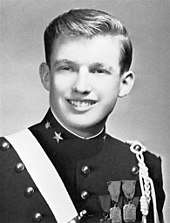 A black-and-white photograph of Donald Trump as a teenager, smiling and wearing a dark pseudo-military uniform with various badges and a light-colored stripe crossing his right shoulder.