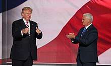 Donald Trump and his running mate for vice president, Mike Pence. They appear to be standing in front of a huge screen with the colors of the American flag displayed on it. Trump is at left, facing toward the viewer and making "thumbs-up" gestures with both hands. Pence is at right, facing toward Trump and clapping.