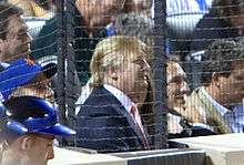 Trump at a baseball game in July 2009. He is wearing a baseball cap and sitting amid a large crowd, behind a protective net.