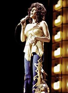 A woman with long brown curly hair, wearing a yellow top and blue pants, singing into a microphone