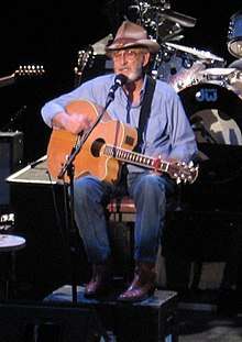 A grey-haired man with a beard wearing a cowboy hat, blue shirt and jeans, playing a guitar and singing into a microphone