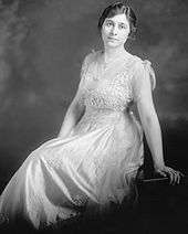 A slightly-built, dark haired woman wearing a white dress and leaning backward onto her left arm
