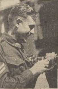 McKenzie is aged 27 and is shown in upper right profile. He is wearing an Australian soldier's uniform with two chevrons just visible at left edge of shot. He is smiling as he looks down at the object he is holding up to chest height.