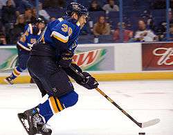 An ice hockey player skates on the ice with his stick and the puck. He is wearing a blue jersey and black pants.