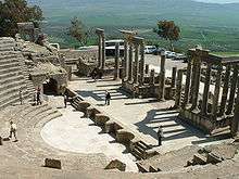 A set of stone seats laid out in a semi-circle to the left overlook a restored stage made up of similar materials to the right. A view of grassy plains precede a wide, mountainous range.