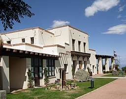 A large white two-story adobe structure with brown trim is fronted by a lawn with stone and metal exhibits, a flagpole, and walkways.