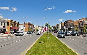 A wide street with two-lane roadways in either direction and older commercial buildings on either side, seen from a grassy median with the American flag flying from a low post in the center