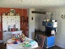 Interior of kitchen: cupboard against wall, table and chairs in foreground, cast-iron stove with pipe leading to chimney