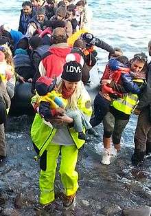 Alison Thompson is dressed in yellow emergency gear and a love cap. She is carrying a Syrian refugee baby to safety.