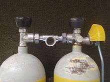 The top of a twin set of steel 7-litre cylinders shows a pair of Dräger vertical spindle taper thread valves with rubber knobs and DIN outlets linked by a single DIN centre-outlet manifold. The left side cylinder has a reserve lever with operating rod and a yellow plastic guard over the lever to reduce the risk of it being inadvertently opened by bumping against the surroundings.