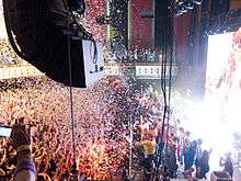 Confetti falling on a packed theater as many people on stage wave to audience.