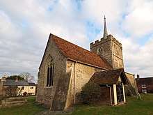 A stone church seen from the south, with a central battlemented tower, the nave with a porch and red tiled roof to the left, and a smaller chancel with a flat copper roof to the right