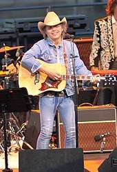A man in a cowboy hat, denim jacket and blue jeans playing a guitar on stage