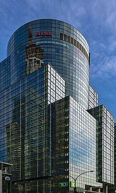 A glass-sided building with mostly rectilinear forms topped by a circular area with the letters "CGI" on them in a red script face, seen from across a signalized urban intersection at its base. Sunlight reflects off its right side, and other nearby buildings and a crane off its left. Behind it is blue sky with some faint altocumulus clouds.