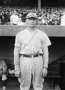 A man in a white baseball uniform with dark pinstripes and a cap with an overlapping "NY" on the front stands in front of a dugout with fans in the stands behind him.