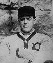 A black-and-white photograph of a man wearing a dark baseball cap and a white sweater with his arms crossed over his chest