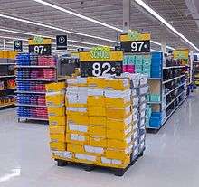 Several people shopping in an area with high shelves on the right stacked with spiral notebooks and other stationery products in open yellow boxes. At the top of the shelves are several blue signs with a small stylized starburst logo in yellow and "Everyday Low Price" in white text, on a red background. Strip fluorescent lights on the ceiling illuminate the scene; a yellow sign hanging from the ceiling has an octagon with "back to school" and text in English and Spanish beneath it. On the left are shelves reaching camera height; a sign in the front bottom says "$9.97".