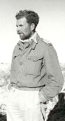 Black and white picture of a man with a short beard wearing a buttoned jacket and light-coloured trousers, facing left with his hands in his pockets
