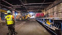 Workers constructing the station's upper concourse, which is located within part of the Grand Central Terminal's Madison Yard. There is construction equipment being stored alongside the walls of the yard.