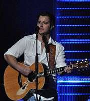 A young man with dark hair wearing a white shirt, playing a guitar and singing into a microphone