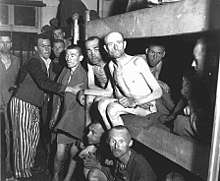 A group of malnutrished prisoners, some of them naked, some dressed in prisoner clothing, fill the interior of one of the barracks, as well as two lower levels of a three-storey bed.