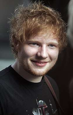 Shot of Ed Sheeran smiling towards the camera, wearing a black T-shirt.