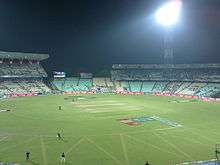 Panoramic view of a cricket ground under floodlights with players occupying the field.