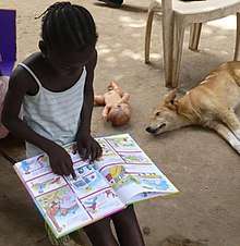 A girl, reading intently. A dog and a doll lie on the ground behind her.