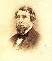 Bust view of bearded man wearing a black jacket, white shirt and collar, black bow tie