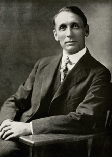 Black and white photograph of a man in an overcoat, sitting in a chair visible from the waist up.  He is clean shaven, has his hair combed to the side.  He wears a calm expression and is looking to the left towards the camera.
