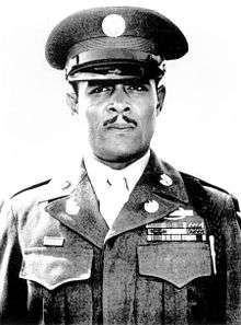 Head and shoulders of a black man, with a carefully trimmed mustache, standing erect and staring directly into the camera. He is wearing a peaked cap and a jacket with three rows of ribbon bars and a pin on the left breast, pins on the lapels, and a braided cord over the left shoulder.