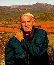 Edward M. Burgess pictured in Namaqualand, South Africa.  Burgess is wearing a dark blue-green jacket and is carrying a camera strapped around his neck (tourist style).  His left arm is positioned across his chest with his left hand on his right shoulder. Burgess is looking directly into the (active) camera with a slight smile.  In the background are numerous mountainous peaks breaking to a pale blue sky at the top of the photo.  At approximately shoulder height the green vegetation in the foot hills gives way to an expanse of orange-red flowers.