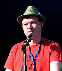 A color photograph of Edwyn Collins, who is speaking into a microphone and peering directly to the left of a microphone.