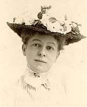 Black-and-white photograph of a woman in a high-necked white dress, wearing a flowered hat