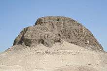 A photo of the pyramid of Senusret II, sitting on a rocky outcropping. Large portions of the pyramid are missing.
