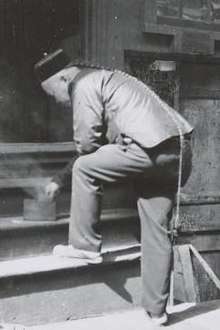 A black-and-white photograph from three-quarter back view of a man wearing a round cap and a long braided queue that reaches to the back of his right knee. His left foot is posed on the first step of a four-step wooden staircase. Bending forward to touch a cylindrical container from which smoke is rising, he is resting his left elbow on his folded left knee.