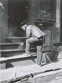 Black and white photo of a Chinese man, who has a very long queue falling along his back, bending over a steaming pan on a step.