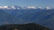 A range of snow-capped mountains. In the foreground is a stretch of forest. In between is an area of lower ground.