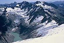 A view looking down on a range of dark mountains. The valley to the left contains a light-colored glacier. There is a small lake at the bottom of the picture.